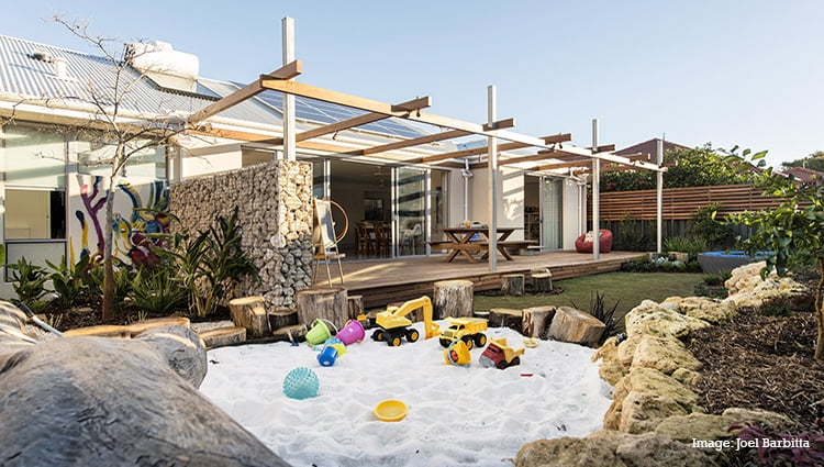 The backyard of a house, with a sandpit filled with toys, a back deck and pergola, and sun shining into the wide open rear French doors, and solar PV and solar hot water panels on the roof.
