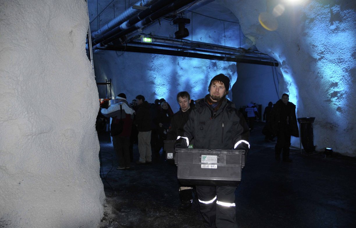 Man in a black waterproof suit and woolen cap carries a black plastic tub in a dimly lit snow tunnel.