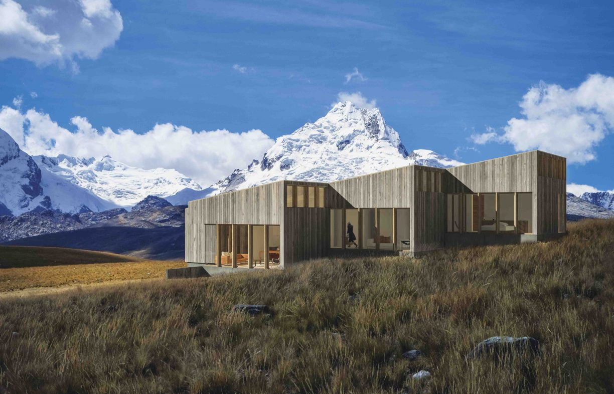 House clad in natural colour, vertical timber panels, located on a sloping grassy landscape with snow-capped mountains in the background.